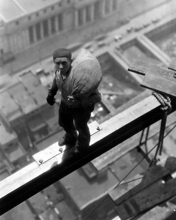 1930. Worker Smoking Cigarette And Carrying Bag Across Shoulders Pauses In The Middle Of Steel Beam High Above City Streets, During Construction Of The Manhattan Company Building At 40 Wall Street, New York City. Photo By Arthur Gerlach