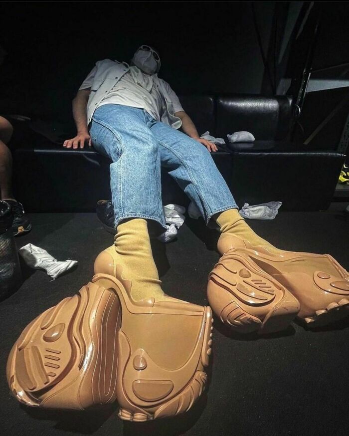 Person in a mask reclining, wearing large, weird shoes and jeans, on a dark couch with scattered socks nearby.