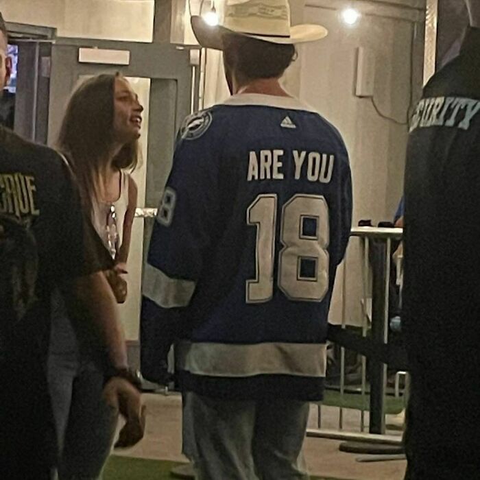 Man in a hockey jersey with "Are You 18" on the back, wearing a cowboy hat, standing in a public area.