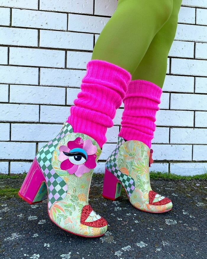 Colorful weird shoes with eye design, pink socks, and green leggings against a white brick background.