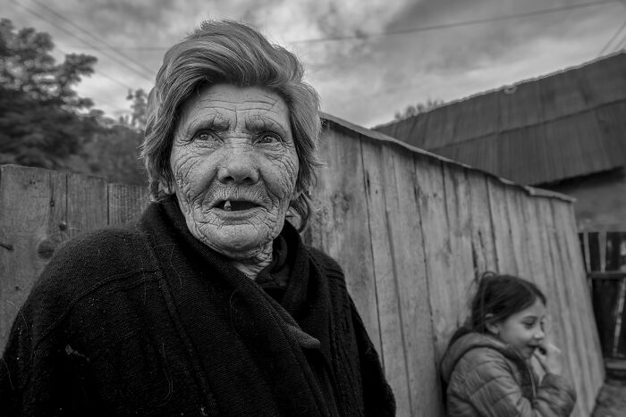 Elderly woman with deep wrinkles wearing dark clothing, standing outdoors with a young girl beside a wooden fence.