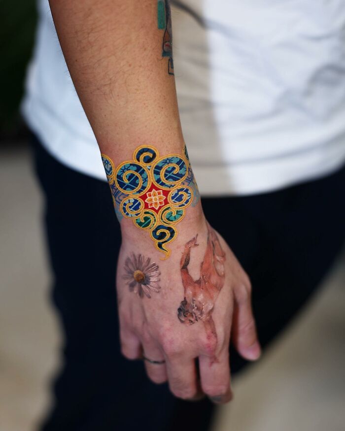Colorful tattoo by Korean artist on a person's wrist and hand, featuring intricate patterns and a floral design.