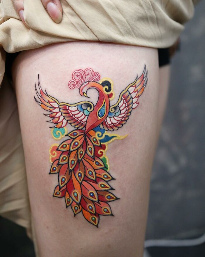 Colorful phoenix tattoo by Korean artist, featuring vibrant reds and blues, on a person's thigh. Mesmerizing tattoo art.