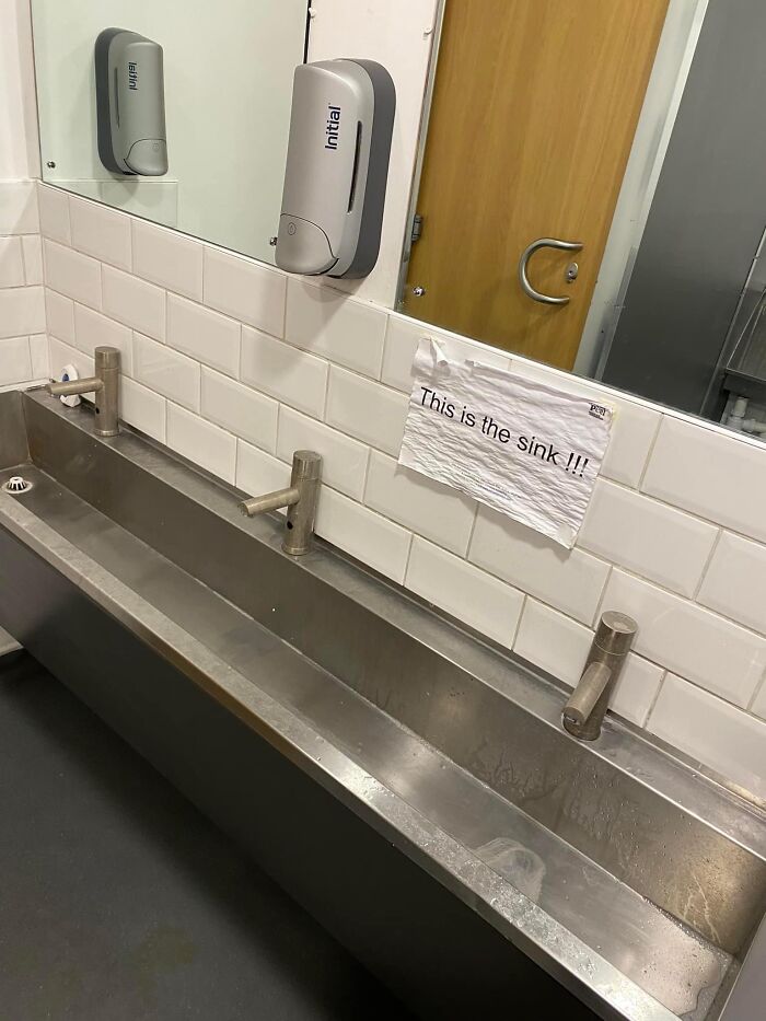 Long stainless steel sink with three faucets and a humorous sign pointing out the sink in a public restroom.