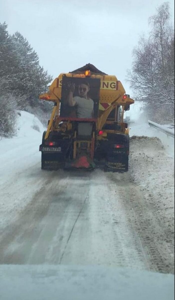 Snow plow truck on icy road with a humorous absurd sign featuring a man in sunglasses making a gesture behind it.