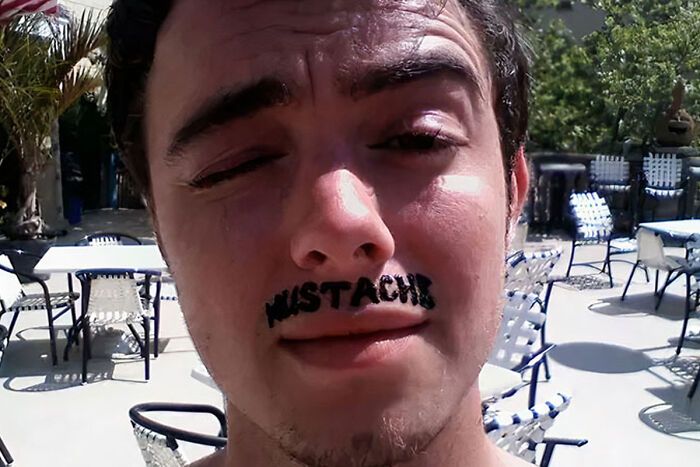 Man with a "mustache" tattoo above his lip, humorously posing outdoors in a sunny setting. 