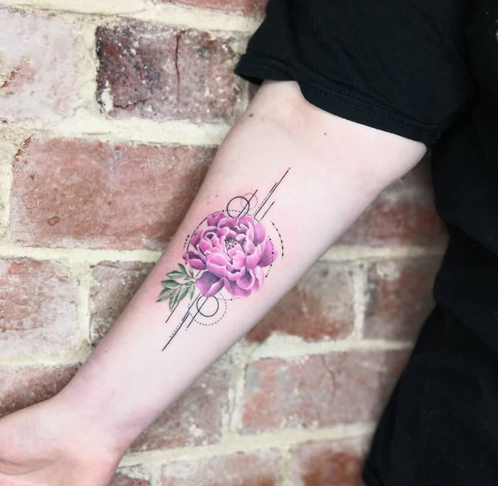 Geometric tattoo featuring a pink peony with circular and linear designs on a forearm against a brick background.