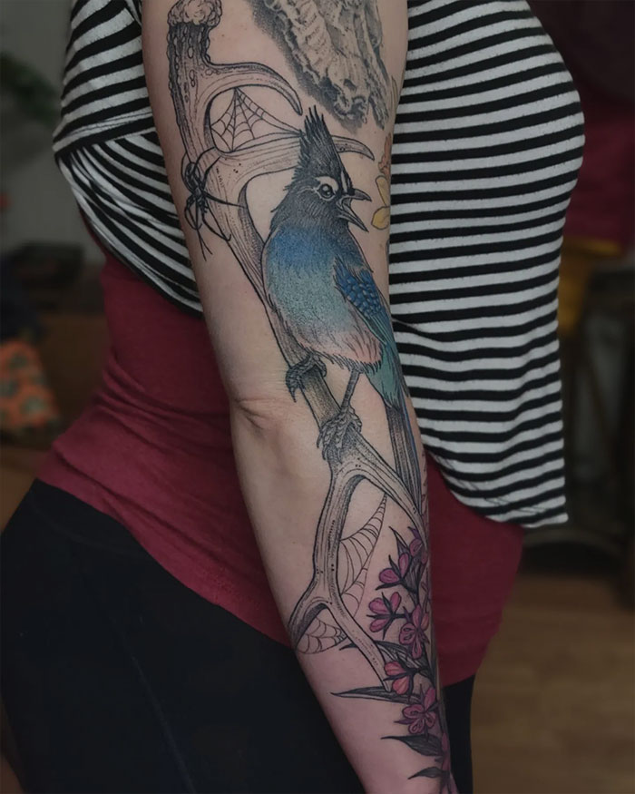 Nature tattoos featuring a vibrant bird, antler, and pink flowers on a person's arm in a striped tank top.