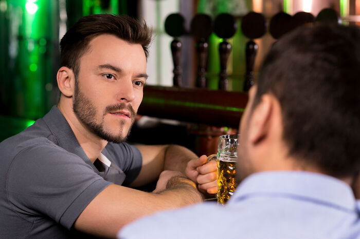 Two men engaged in a serious conversation at a bar, discussing tattoos over drinks.