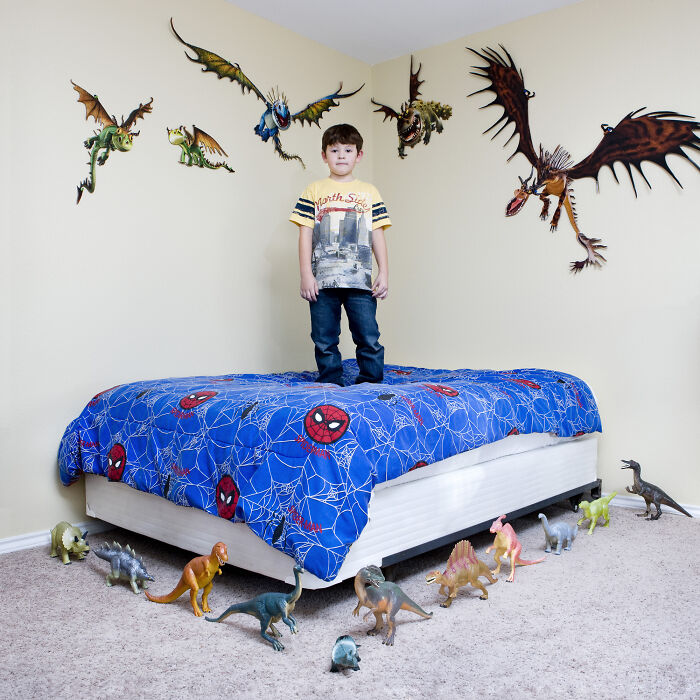 Young boy standing on bed surrounded by dinosaur toys in a room, showcasing toy collections from the series Toy Stories.
