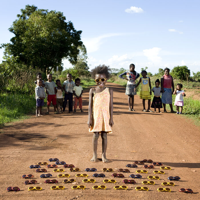 Child from the Toy Stories series standing on a dirt road with colorful sunglasses arranged as a toy collection around her.