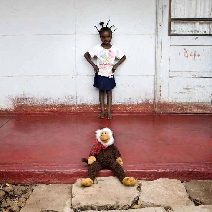 Young child with a stuffed monkey toy on a red porch, part of a series showcasing children’s toy collections worldwide.