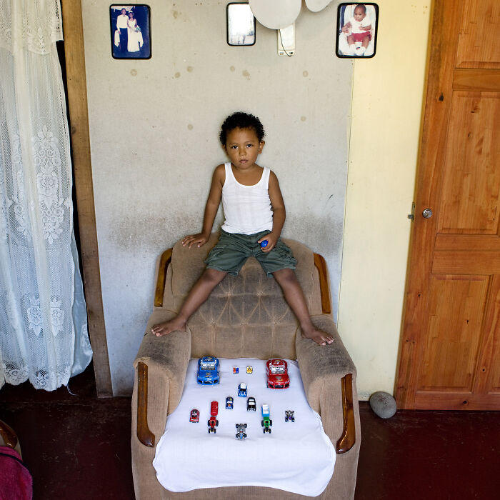 Young boy from the Toy Stories series sits on a chair showcasing his collection of toy cars in a modest home.