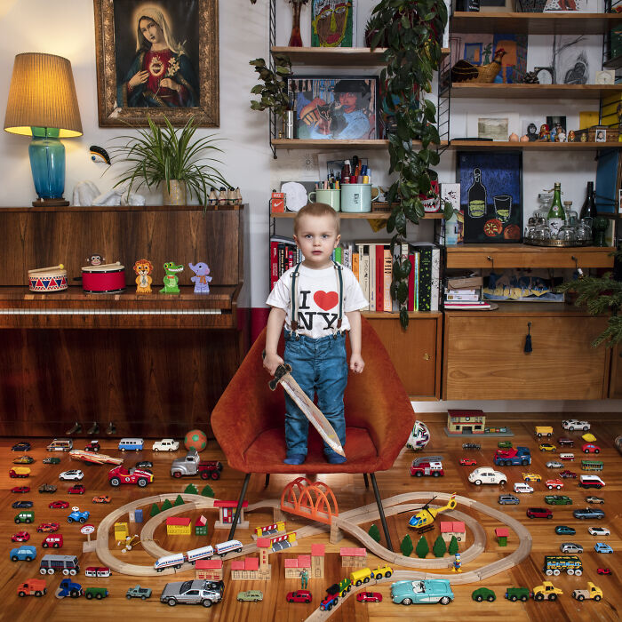 Young boy standing with a toy sword surrounded by an extensive toy collection featuring cars and a wooden train set.