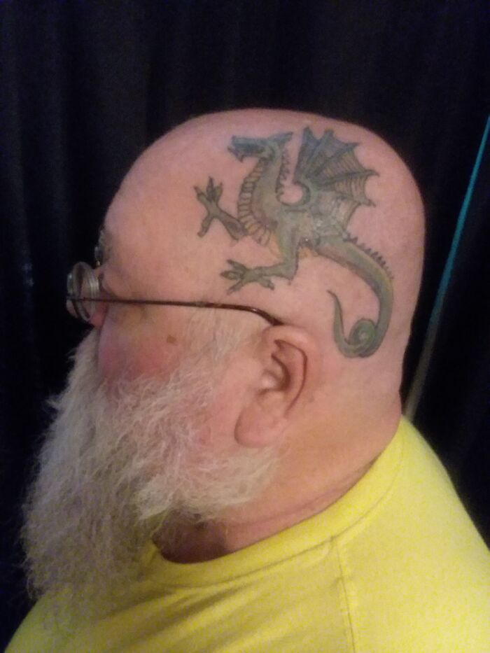 Bald man with a dragon tattoo on his scalp, wearing glasses and a yellow shirt.