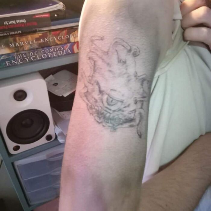 Bad tattoo of an unclear design on a person's arm, with comic book encyclopedias in the background.