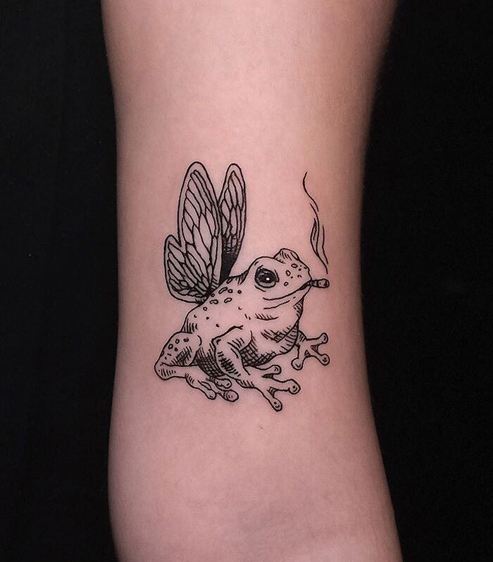 Animal tattoo of a frog with insect wings and a cigarette on an arm.