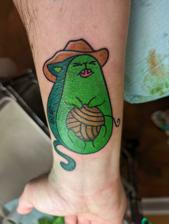 Funny tattoo of a green cat wearing a cowboy hat, holding yarn on a forearm.