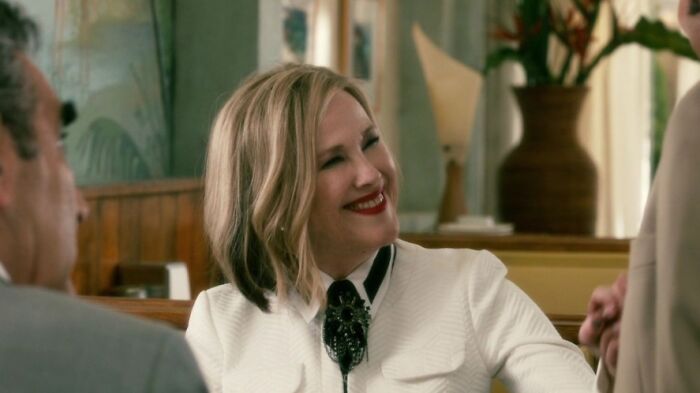 Moira Rose in a white top is smiling and sitting in a cafe