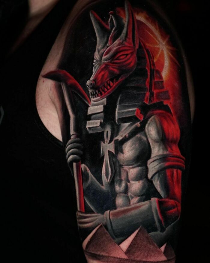 Realistic tattoo of an ancient Egyptian god with Anubis features and vibrant red accents on a shoulder.