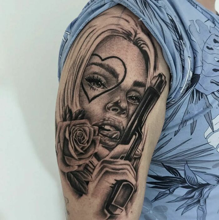 Super realistic tattoo of a woman with a heart face tattoo and a rose, holding a gun, on a person's arm.