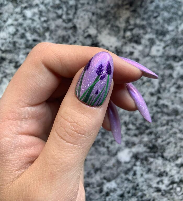 Purple floral nail art on a hand, showcasing a trendy manicure design on a stone background.