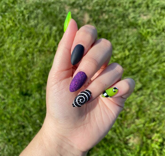 Colorful nail art design featuring black, purple glitter, spiral black-and-white, and neon green patterns on a hand.