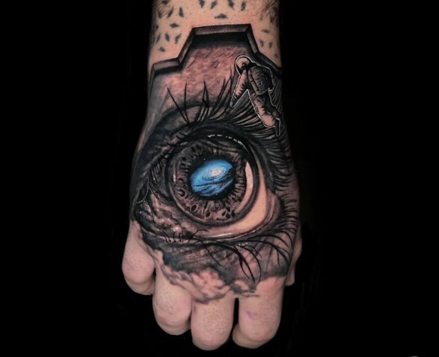 Realistic tattoo of a detailed eye on a hand, featuring intricate shading and a vibrant blue iris design.