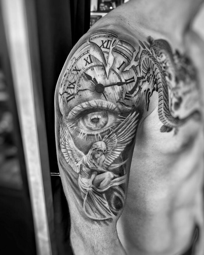 Realistic tattoo of an eye and angel on a man's upper arm, featuring detailed clock elements, in black and gray tones.