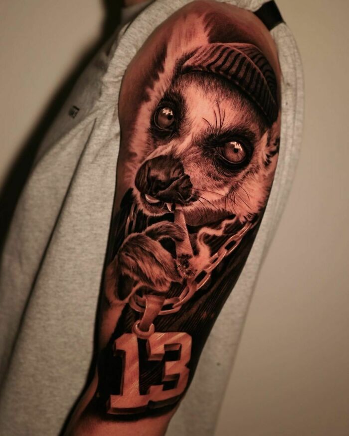Realistic tattoo of a lemur with a number 13, styled with a chain and hat on a person's arm.