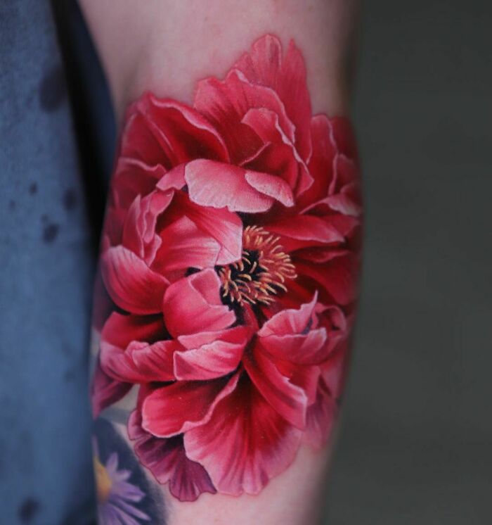 Realistic tattoo of a vibrant red flower on an arm, showcasing intricate details and vivid colors.