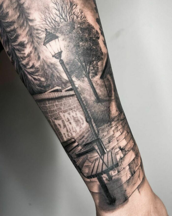 A super realistic tattoo depicting a street scene with a lamp post, trees, and a pathway on a person's forearm.