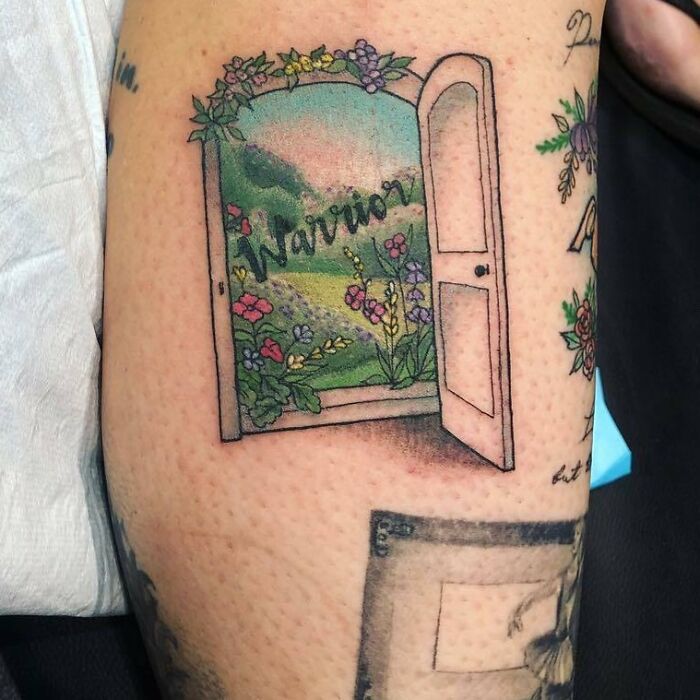 Word tattoo of "Warrior" inside an open door with floral landscape on arm.