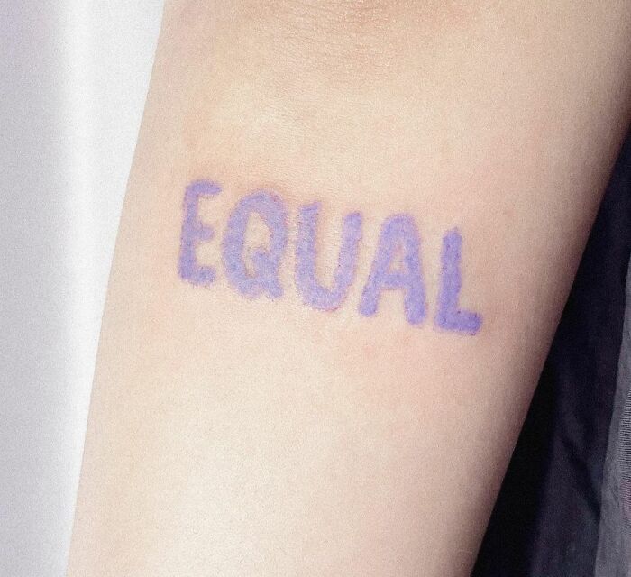 Tattoo of the word "EQUAL" in purple ink on forearm, representing creative word tattoos for ink inspiration.