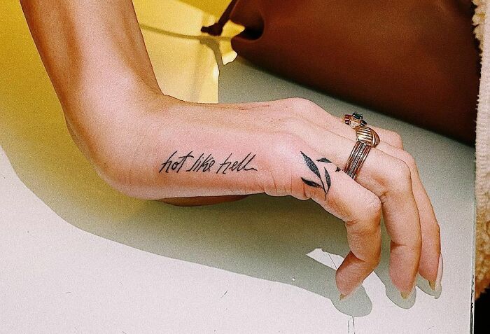 Hand with creative word tattoo reading "hot like hell," adorned with rings, showcasing inkspiration.