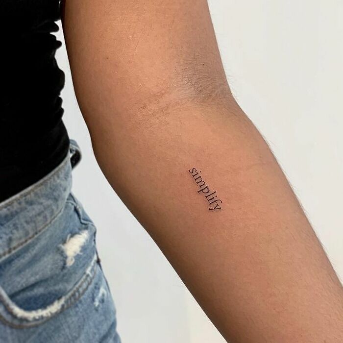 Arm with "simplify" word tattoo in minimalist style, jeans visible, reflecting creative inkspiration.