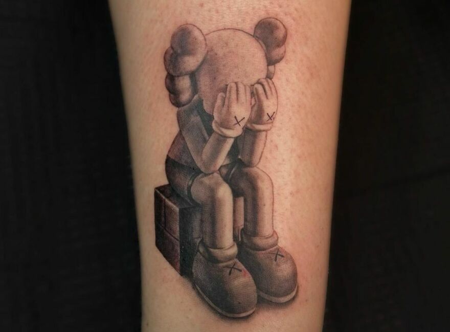 Realistic tattoo of a kaw figure sitting on a box, covering face with hands, showcasing detailed artistry.