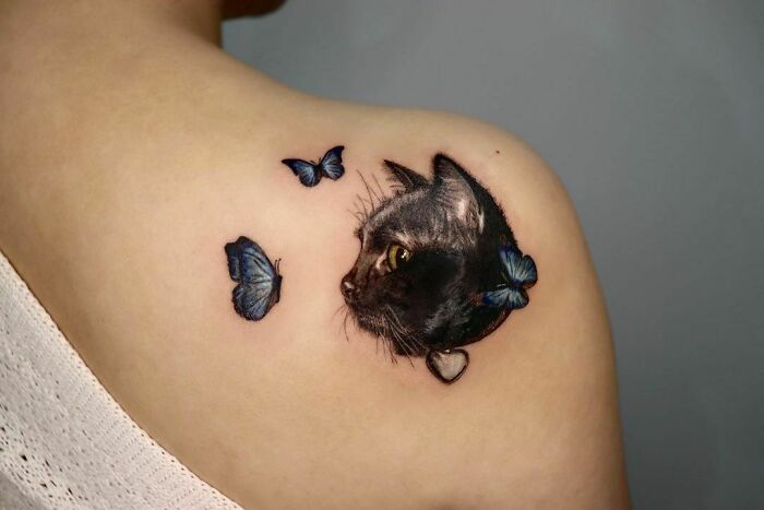 Realistic tattoo of a cat with butterflies on a person's shoulder.