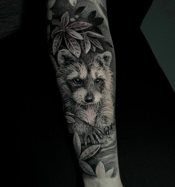 Realistic tattoo of a raccoon with detailed fur and leaves on a forearm, showcasing museum-level art.