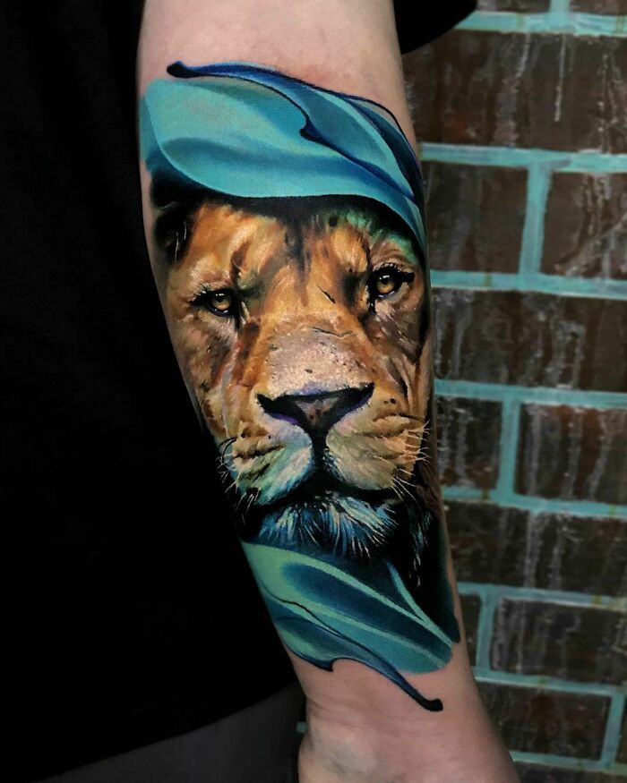 Realistic lion tattoo on forearm, showcasing intricate details and vibrant colors, set against a brick wall background.
