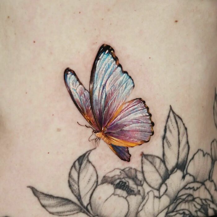 Realistic tattoo of a colorful butterfly on skin, surrounded by intricate black and white floral design.