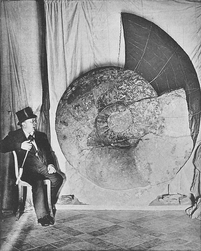 Black and white photo of a man in a top hat seated next to a giant fossil, showcasing nature was metal.