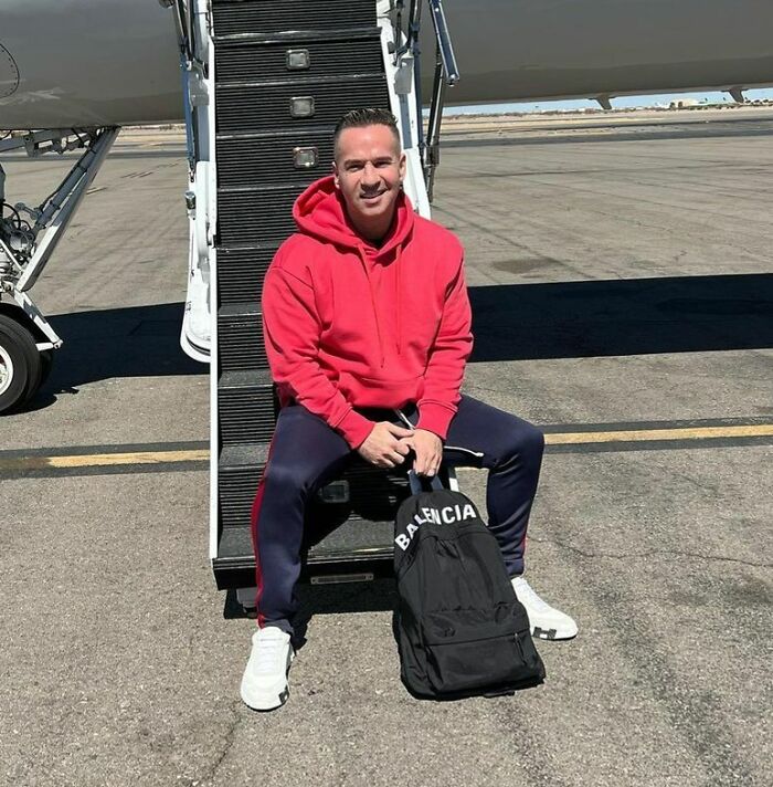 Celebrity in red hoodie sitting on plane stairs with a Balenciaga bag, outdoors.