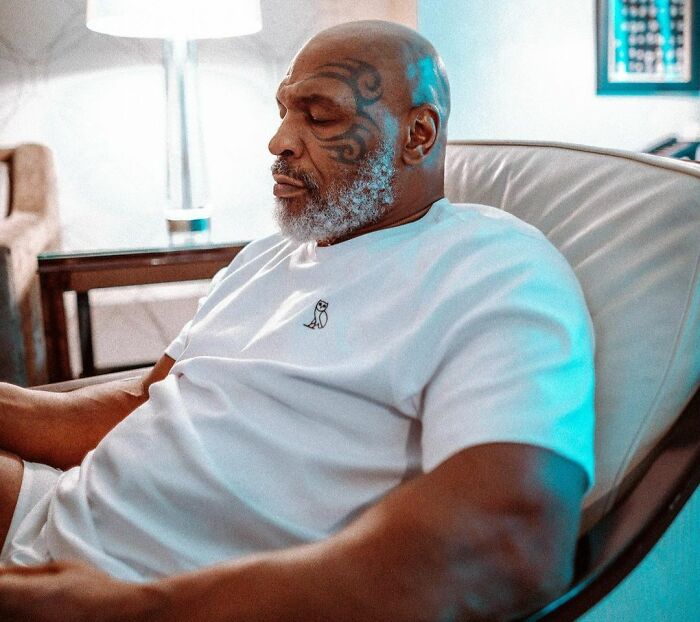 Celebrity relaxing in a chair, wearing a white shirt, showcasing facial tattoo.