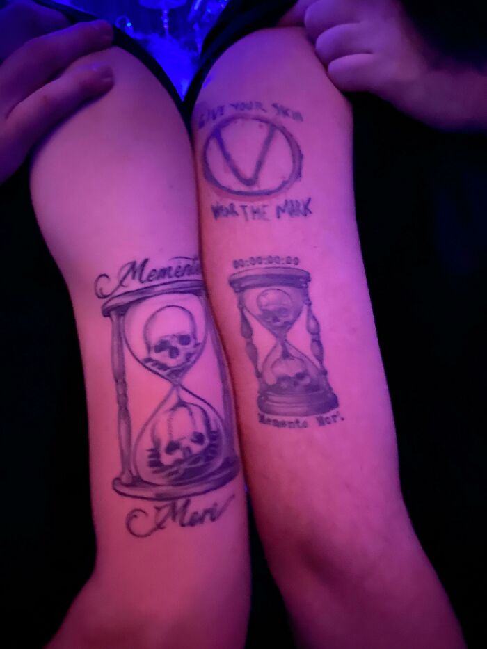 Matching tattoos with skulls and hourglasses on arms, perfectly aligned in a low-light setting.