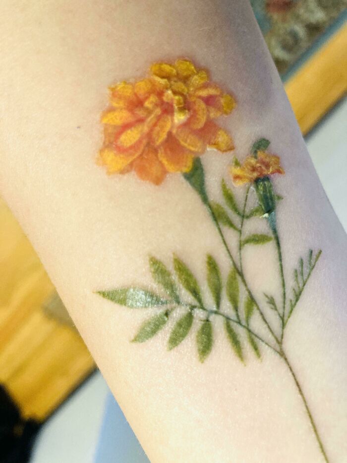 Tattoo of a marigold flower on an arm, matching the natural look of surrounding foliage.
