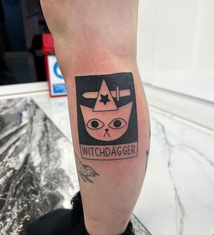 Calf tattoo featuring a cat with a hat and dagger, labeled "Witchdagger," showcasing cool tattoo art.
