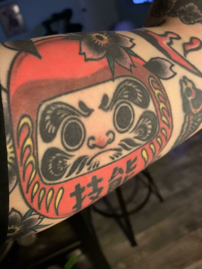 Tattoo featuring a Daruma doll design on an arm, showcasing intricate details and bold colors.
