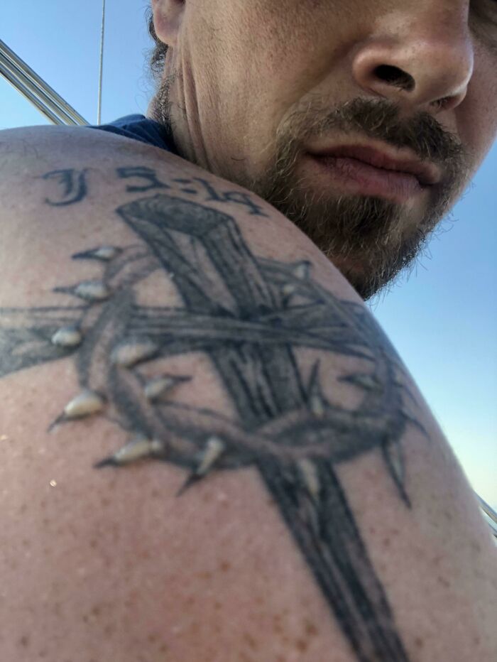 Man with a tattoo of a cross and thorn crown on shoulder, perfectly matching the moment.