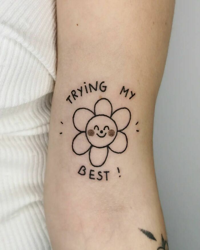 Smiling flower self-love tattoo with text "Trying my best" on an arm.